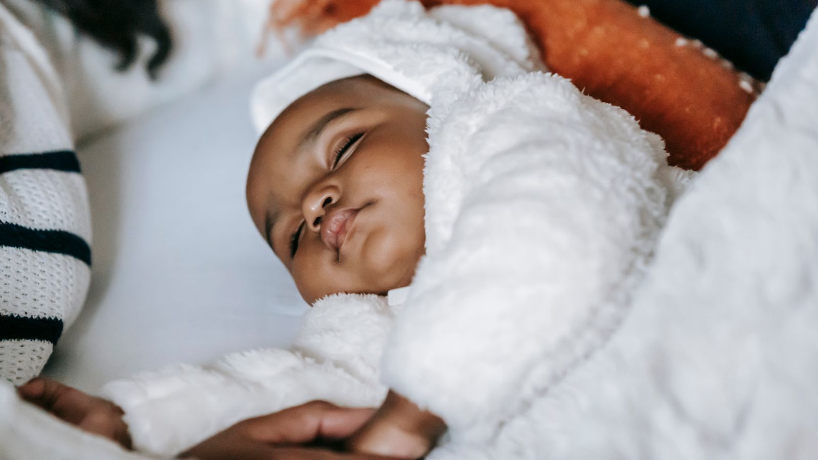 Een ouder schrijft met de hand in een notitieboek aan een houten tafel, een slapende baby zichtbaar op de achtergrond in zacht ochtendlicht